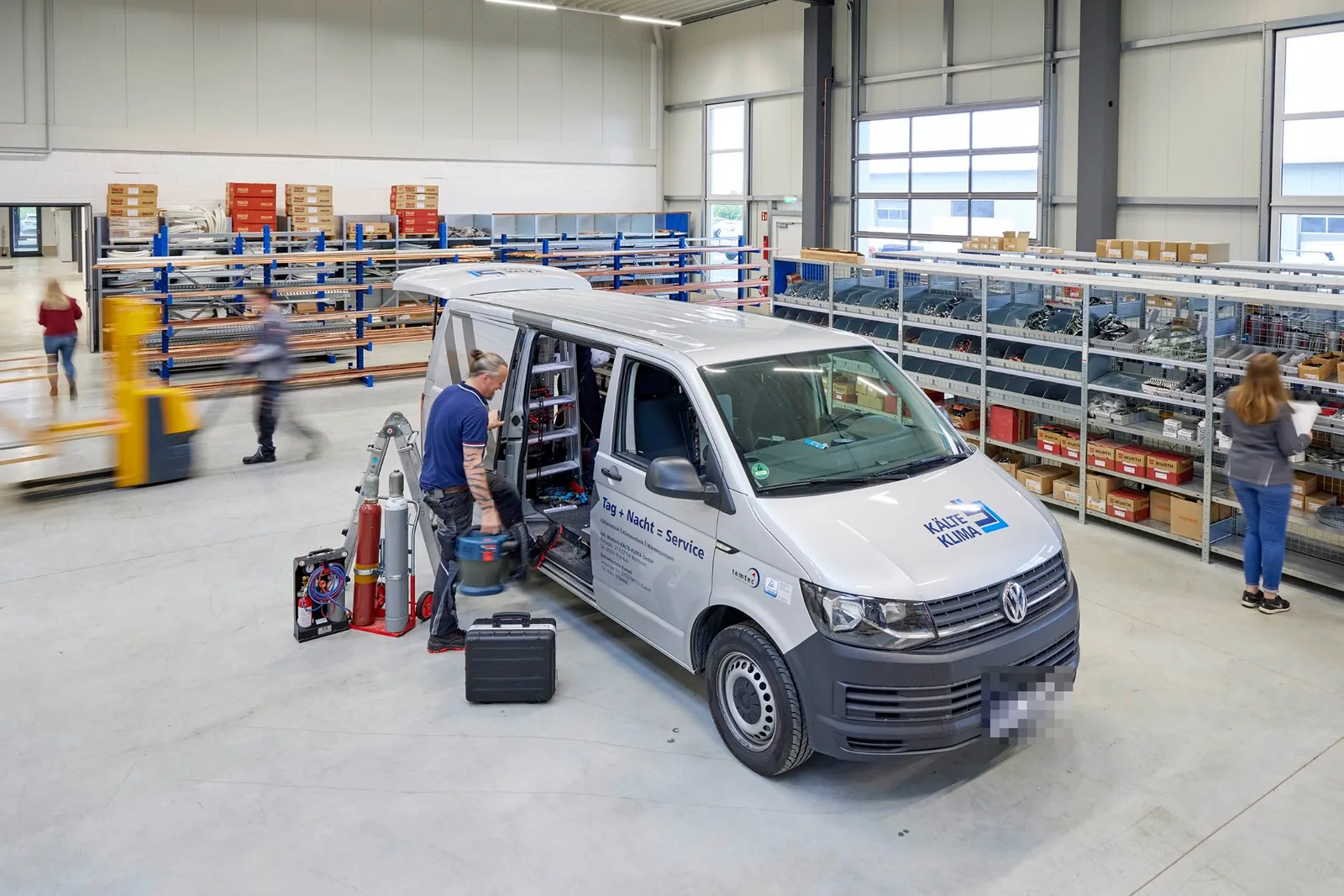 Monteur am Servicewagen mit der Aufschrift „Tag + Nacht = Service“ in der Werkstatt- und Lagerhalle, im Hintergrund Regale mit Material