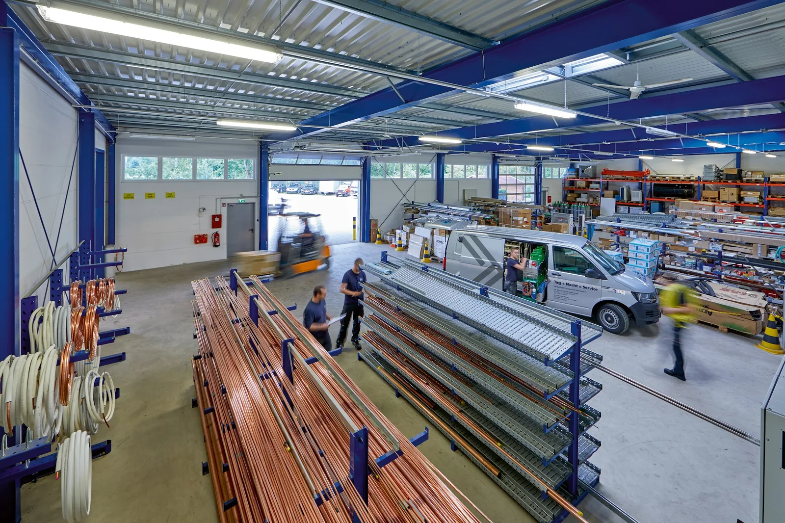 Lagerhalle mit blauen Stahlträgern, Regalen voller Kupferrohre, zwei Mitarbeitern und einem Servicewagen