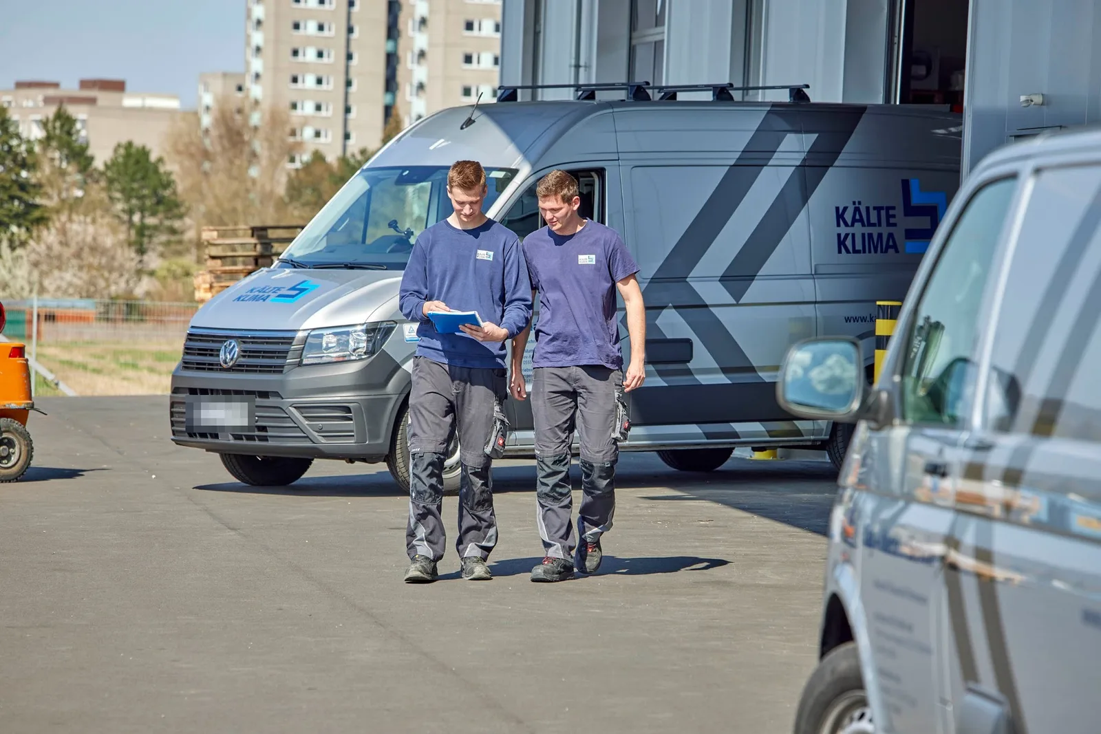 Zwei Monteure in KÄLTE-KLIMA-Arbeitskleidung gehen mit einer Mappe über den Hof, dahinter ein beschrifteter Transporter