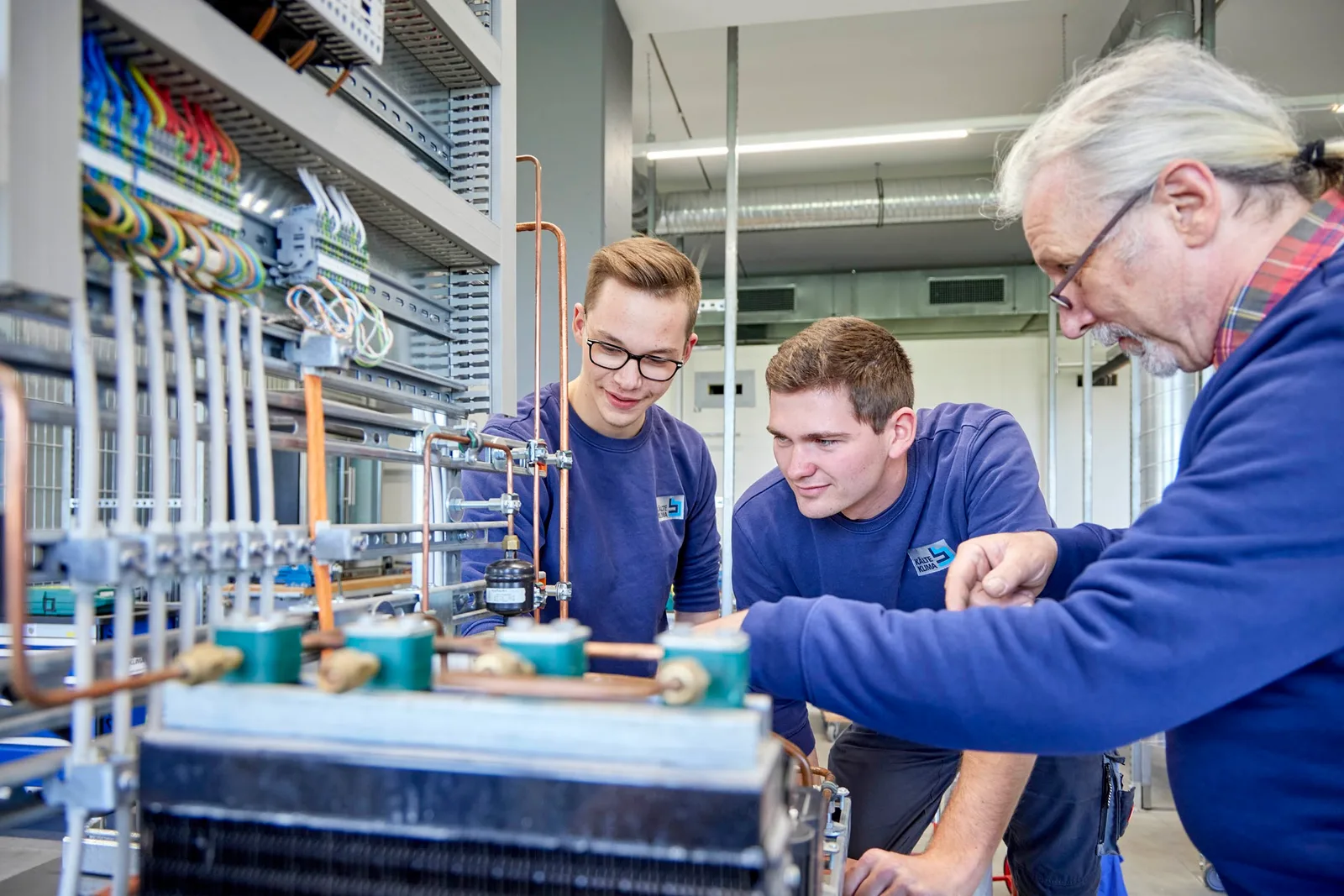 Ein Ausbilder zeigt zwei jungen Mitarbeitern in KÄLTE-KLIMA-Pullovern etwas an einem Übungsstand mit Kupferleitungen und Magnetventilen