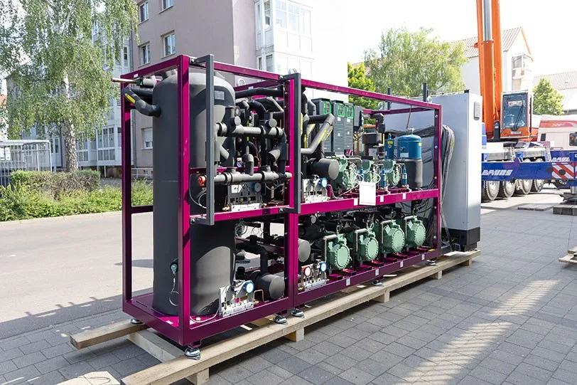 Neue CO2-Verbundkälteanlage im violetten Rahmen bei der Anlieferung vor einem Supermarkt, dahinter ein Kran